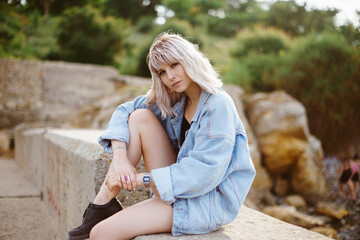 Blonde hipster girl with blonde hair in jeans jacket sitting outdoors on the rocks.