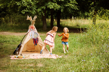 Funny blonde girls playing next to wigwam in the forest. Children games, leisure time. © Natalia