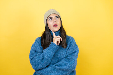 Young beautiful woman wearing blue casual sweater and wool hat serious face thinking about question with hand on chin, thoughtful about confusing idea