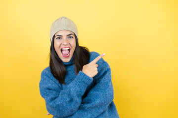 Young beautiful woman wearing blue casual sweater and wool hat smiling and pointing with hand and finger to the side