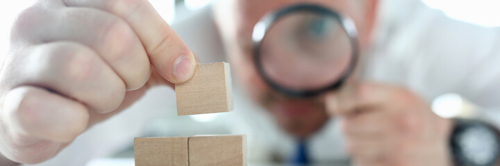 Close-up of man looking on wooden cubes stack through magnifying glass. Concentrated person building pyramid on working table. Business strategy and smart decision concept - obrazy, fototapety, plakaty