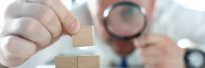 Poster Close-up of man looking on wooden cubes stack through magnifying glass. Concentrated person building pyramid on working table. Business strategy and smart decision concept © H_Ko