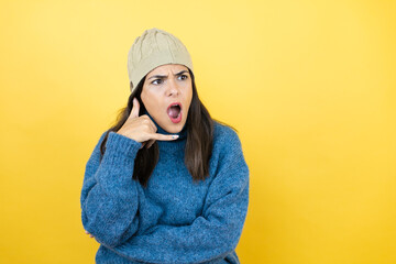 Young beautiful woman wearing blue casual sweater and wool hat confused doing phone gesture with hand and fingers like talking on the telephone
