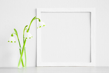 Fresh snowdrops in glass vase on table at light gray blue wall. First messengers of spring. Empty place for inspirational, emotional, sentimental text, quote or sayings in white frame. Closeup.