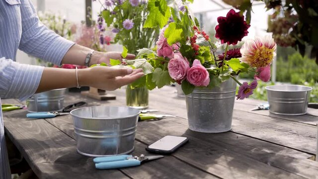 Unrecognizable woman in light blue dress puts bouquets with colorful flowers in vases on long wooden table preparing for masterclass in greenhouse