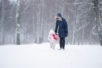 Little daughter in overalls and young adult mother walking on white first snow at park. Spending time together in beautiful winter day. Enjoying peaceful stroll. Front view.