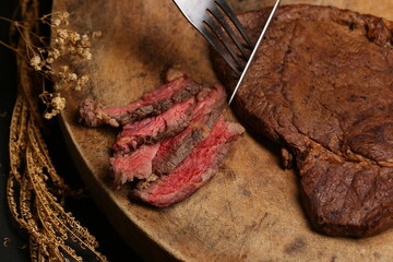 Juicy medium Beef Rib Eye steak slices in pan on wooden board with fork and knife herbs spices and salt.