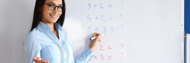 Portrait of sweet brunette school teacher explaining information on white board. Smiling attractive lady wearing glasses. Mathematics lesson and education system concept