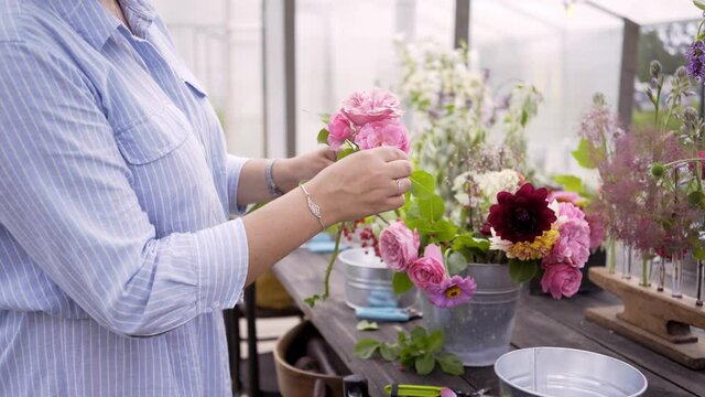 Faceless woman wearing light blue dress and straw hat arranges bouquet with fresh colorful rose flowers at table in modern greenhouse on summer day