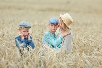 Mom in a blue dress and hat plays with her sons in the wheat field caps. Family summer vacation in the village.
