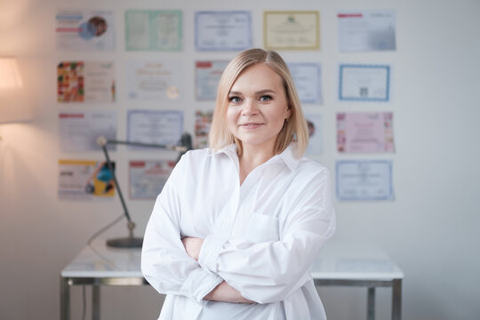 Portrait of blonde woman doctor in a beauty salon in white uniform, beautician looking at the camera and smiling, master of permanent makeup stands on a background of certificates, selective focus