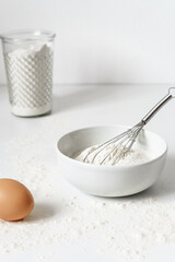 Fototapeta na wymiar Mehl in einer Schüssel, ein Schneebesen und ein braunes rohes Ei auf einem weißen Küchentisch. Vorbereitung, Frühstück, backen.