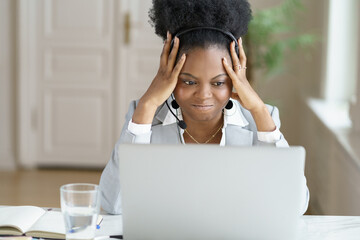 Mad angry annoyed afro businesswoman screaming, wear headphones, talking to a bad customer looking at screen laptop in office. Office employee confused by message of a disgruntled buyer, touching head - obrazy, fototapety, plakaty