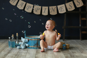 Naked kid sitting on a wooden floor playing with soap bubbles. Studio shot, dark background. Concept of children and bathing