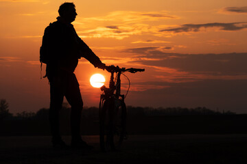 Obraz premium The silhouette of a cyclist against the background of the sun and the beautiful sky. The outline of a man standing next to his bicycle against the background of the sunset.