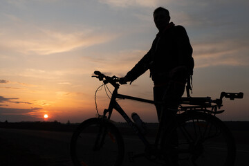 Obraz premium The silhouette of a cyclist against the background of the sun and the beautiful sky. The outline of a man standing next to his bicycle against the background of the sunset.