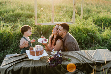 Young parents and little son eat cake together in a field at sunset.