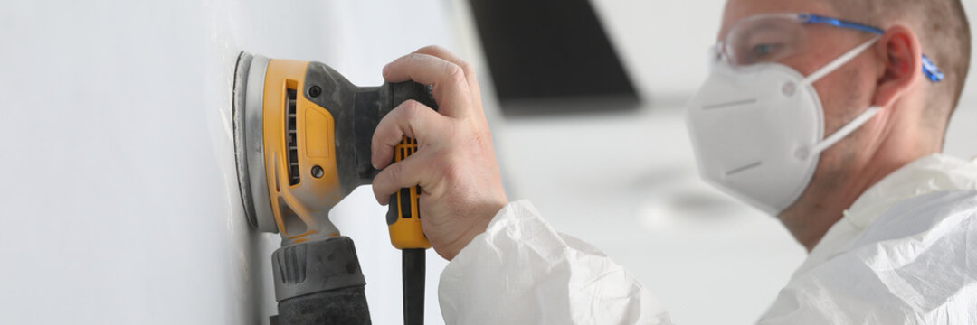 Close-up of professional worker wearing white protective face mask and working with yellow sander machine. Qualified man polish wall in apartment. Construction site and renovation concept