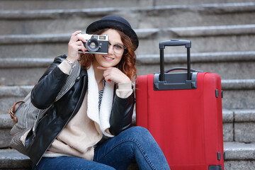 giovane turista in abiti invernali seduta nei gradini con il trolley vicino, fa una fotografia con...