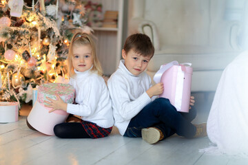 nice little girl 3 years old blond boy and 5 years in white knit sweater sitting on a white wooden floor with gifts in pink boxes under decorated Christmas tree