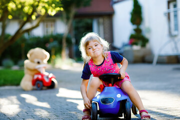 Little adorable toddler girl driving toy car and having fun with playing with plush toy bear, outdoors. Gorgeous happy healthy child enjoying warm summer day. Smiling stunning kid in gaden