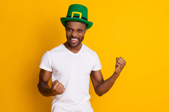 Portrait of delighted afro american guy win saint patrick contest raise fists scream yes wear green hat white clothes isolated over bright shine color background
