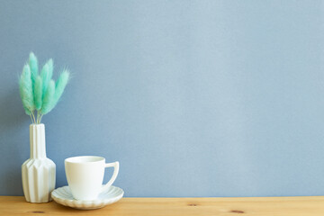 Coffee cup with vase of Lagurus ovatus dry grass on wooden table. Blue background, copy space