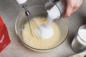 Female hand adds sugar to a transparent bowl with whipped eggs, uses a mixer. Ingredients for baking cake. Cooking set. Glass of flour, pack of sugar. Culinary class in a kitchen.