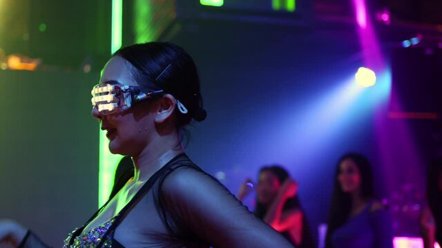 Confidence Asian woman wear LED glowing glasses dancing to the music played by dj with multi-color illuminated neon night lights at night club. Group female enjoy nightlife and having fun dance party.