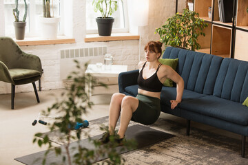 Obraz premium Squats workout with sofa. Young woman exercising fitness, aerobic, yoga at home, sporty lifestyle and home gym. Getting active during lockdown, quarantine. Healthcare, movement, wellness concept.