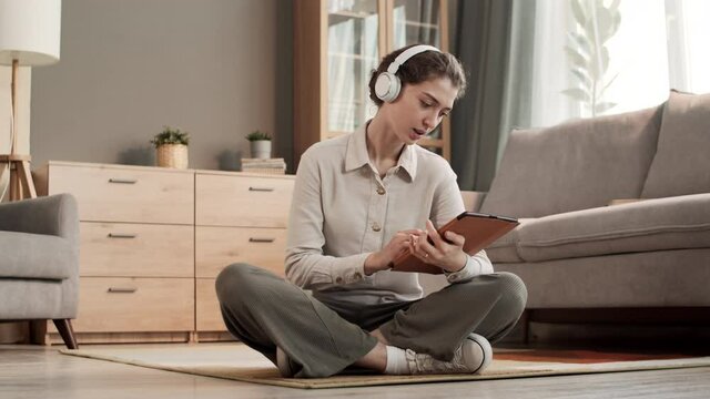 Full shot of young Caucasian woman wearing wireless headphones, sitting with legs crossed on floor in living room, using tablet computer, looking pleased and surprised