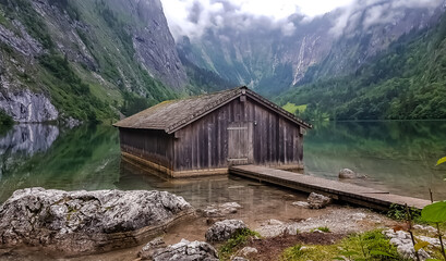 Fototapeta na wymiar Romantic wooden boathouse at the lake Obersee next to lake Königssee in the Bavarian alps. In the background flows Germanys highest waterfall Röthbachfall.