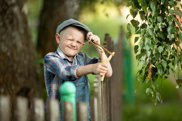 Funny little boy a bully in a cap and plaid shirt standing at a wooden fence shooting a slingshot....