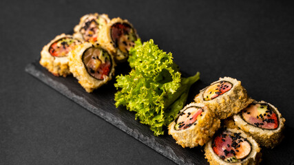 Obraz na płótnie Canvas Custom sushi roll in tempura with nori, fresh salmon, tuna, avocado, masago caviar, drizzled with pineapple sauce with salad pouring as decoration on a black plate on a black table and background.