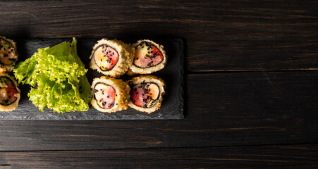 Custom sushi roll in tempura with nori, fresh salmon, tuna, avocado, masago caviar, drizzled with pineapple sauce with salad pouring as decoration on a black plate on a wooden table and background.