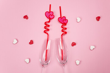 Two champagne glasses with red plastic drinking straws with hearts on a pink background.