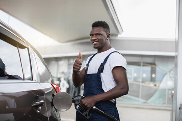 Fototapeta na wymiar Close up portrait of a happy young African guy, professional gas station worker in uniform, smiling and showing thumb up to camera, while refueling modern car