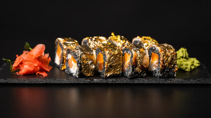 Custom sushi roll with nori, cuttlefish ink, fresh salmon, cream cheese, pepper pumpkin, eel, unagi sauce and edible gold leaf with red ginger, wasabi on a square plate on black table and background.