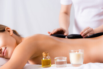 Obraz na płótnie Canvas aromatic candle and bottle with oil near young woman getting hot stone massage on blurred background