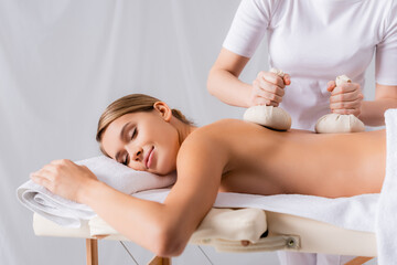 woman massaging young pleased client with herbal bags in spa salon