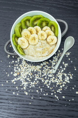 Oatmeal porridge with bananas, kiwi in in a bowl on a dark background, top view. Healthy breakfast. Vertical photo
