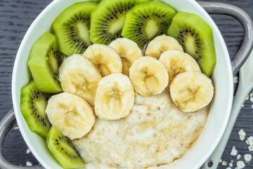 Oatmeal porridge with bananas, kiwi in a bowl on a dark background close-up. Healthy breakfast.