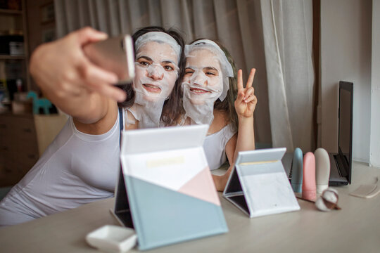 Beautiful girl and her mother using gadgets during spa procedure, mom and daughter chatting via internet with smooth mask on face, wellness, relaxation and beauty salon at home, indoor lifestyle