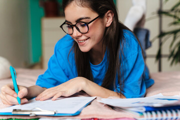 Happy brunette girl in eyeglasses doing homework while lying on bed