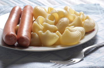Close-up of meat sausages with boiled pasta in a plate and a fork on a napkin
