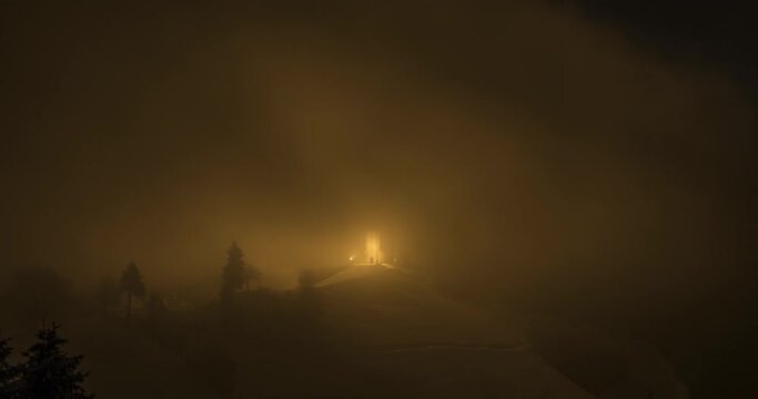 Time lapse view of fog moving over Jamnik church at night. Light illuminating inversion clouds with yellow color. Famous landmark in Slovenia. Low visibility in dense fog. Zoom out