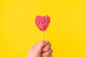 Handholds red heart-shaped lollipop candy isolated on an empty colorful yellow background. Symbol of love for Happy Women's, Valentine's Day, Birthday greeting romantic postcard