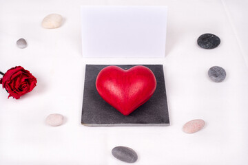 Top view on a red love heart presented as a jewel on a square slate, with a white and blank gift card behind, and stones arranged around and a red rose. Love and valentine's day 