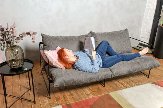 Young woman with red hair is lying on couch and reading book. Quiet leisure at home with book in cozy interior. Concept: love of literature, self-study. - Powered by Adobe