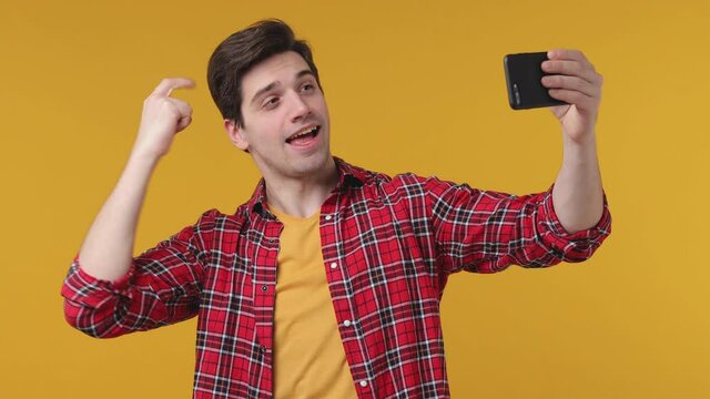 Funny young man 20s in red checkered shirt posing isolated on yellow background studio. People lifestyle concept. Doing selfie shot on mobile phone point finger himself showing thumb up biceps mucles
